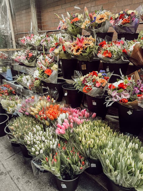 A wide view of a flower shop display featuring a variety of freshly cut flowers arranged in black buckets and wrapped bouquets. In the foreground, there are clusters of pink, white, and orange tulips,