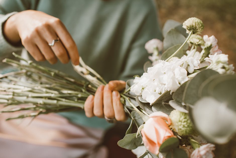 A professional florist from flowerdeliverybarbican.co.uk is arranging a bouquet of fresh flowers, including white hydrangeas, pale pink roses, and green foliage, with stems wrapped in natural twine. T