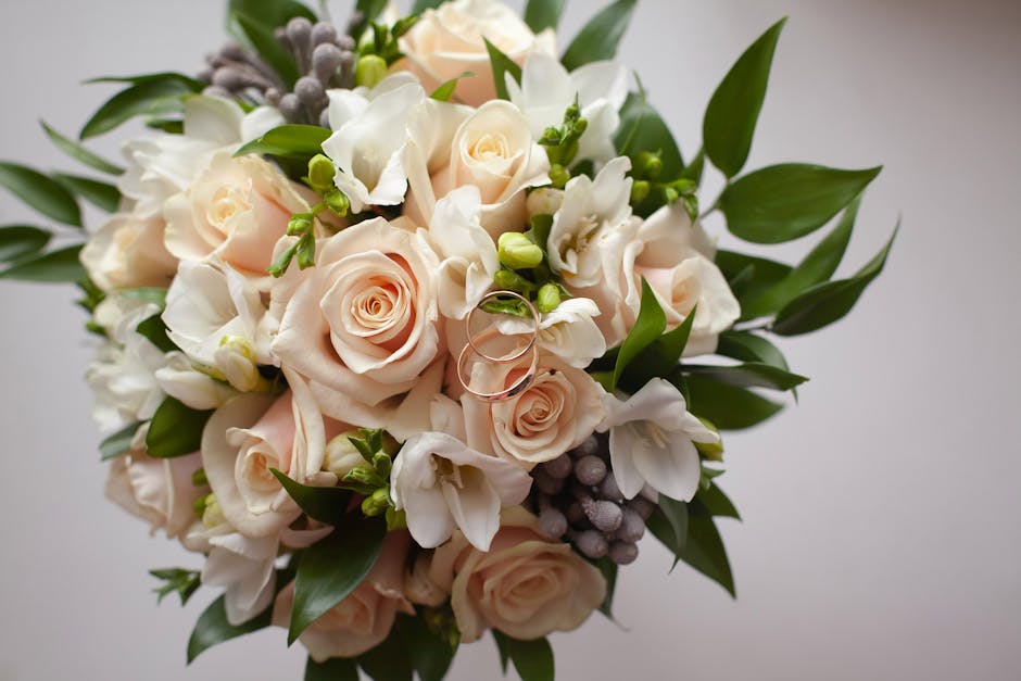 A close-up view of a floral bouquet arranged with pale peach roses, white freesias, green foliage, and clusters of small purple berries, elegantly wrapped with light-colored paper and tied with a ribb