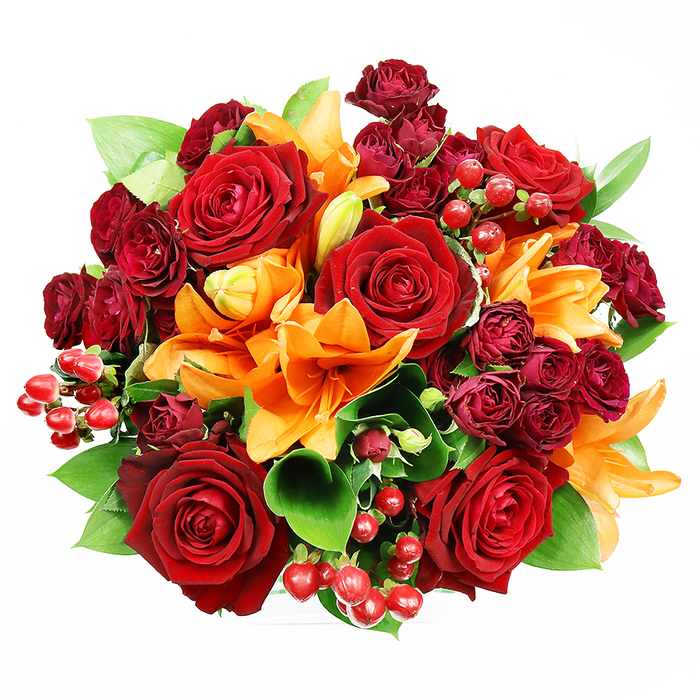 Captured from above against a pristine white backdrop, this opulent, circular bouquet presents a carefully layered study in warm tones and tactile contrasts. At its core several large, fully bloomed deep red roses anchor the design with velvety petals that unfurl in classic spirals, evoking timeless romance and depth. Scattered among them, bright orange Asiatic lilies introduce dramatic trumpet silhouettes and visible stamens-some blooms fully expanded, others in the graceful tension of an opening bud-creating a dynamic rhythm across the surface. Complementing these are plentiful clusters of small, dark red spray roses, their tightly packed petals forming miniature rosettes that add density and a jewel-like richness to the palette. Intermittent clusters of glossy, spherical Hypericum berries in ruby tones provide crisp, polished highlights and tactile diversity, their smooth skins contrasting with the roses' soft matte. Fresh green foliage-broad ruscus or salal leaves-emerges between the flowers to offer cooling relief and structural support, their sheen and shape accentuating the bouquet's rounded silhouette. The lighting is even and soft, isolating the composition on white so every texture and color shift reads clearly; the overall impression is sumptuous yet carefully balanced. The scent is imagined as a layered perfume: the deep, warm note of red roses tempered by the lily's bright, spicy-citrus lift. This bouquet suits anniversaries, romantic gestures, or autumnal celebrations and would make a striking centrepiece in a Barbican townhouse or on a table near London Wall. It speaks to considered craftsmanship-the kind of arrangement a seasoned local florist would select for meaningful moments.